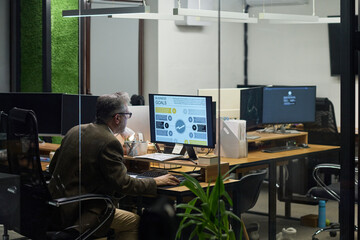 Fototapeta na wymiar Middle aged Caucasian man working at desk using desktop computer with business analytics displayed on screen in modern office environment, sitting alone in workspace