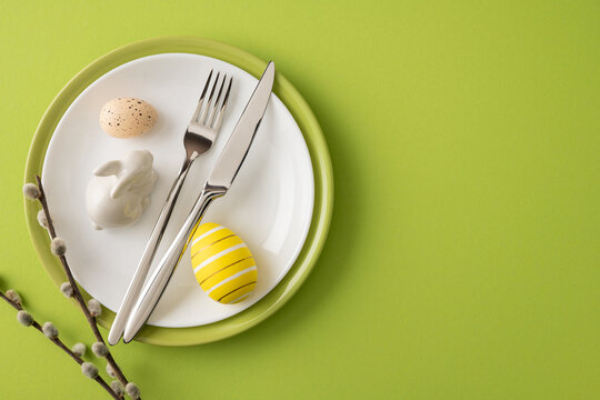 Happy Easter table setting featuring a striped yellow egg, speckled egg, ceramic bunny figurine, silver cutlery and willow sprigs on a green plate and background