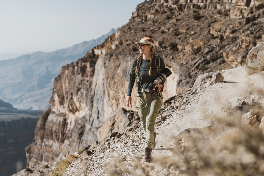 Woman carrying camera hikes along dramatic canyon edge, Oman