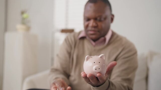 Average citizen saving money for retirement or purchase, holding piggy bank. Portrait of serious african american man, black man counting incomes and putting money into funny ceramic money-box