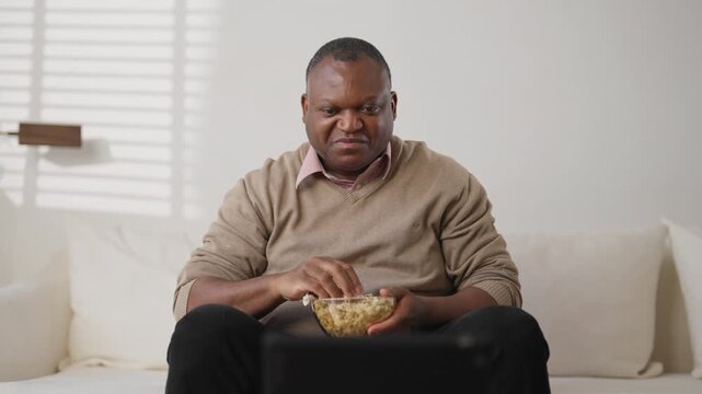 Lazy weekend at home, mature black man watching TV and eating popcorn, portrait. Entertainment and leisure in evening at home, middle-aged african american male person enjoying TV show or sport match