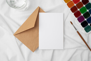 Fototapeta premium Blank Greeting Card Mockup with Envelope, Watercolor Paint Palette and Brush on White Background