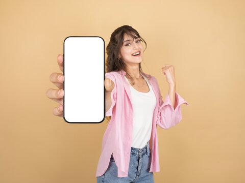 Smiling young woman holding smartphone with blank screen toward camera while celebrating success, posing against beige studio background with large copy space for advertising. Advertisement concept.