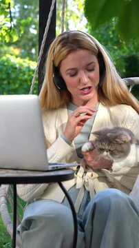 Middle aged woman working remotely with laptop and cat on her lap on terrace. Warm light, cozy lifestyle, family emotions, peaceful outdoor home office vibe.