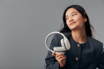 Young woman in dark jacket holding white headphones looking sideways with slight smile against gray...