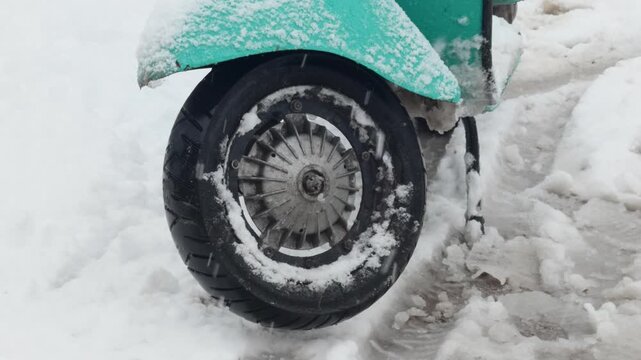 Detailed close-up of a vintage scooter front wheel coated with snow, parked in slushy winter conditions with falling snow.