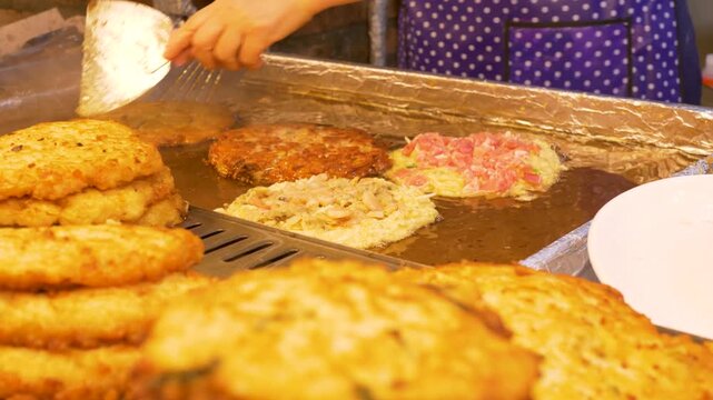 Slow motion close up of local vendor using cooking utensils to flip Bindaetteok seafood pancake on hot grill frying in oil at Gwangjang night street markets in Seoul city South Korea Asia recipe