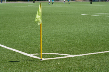 A yellow corner flag stands on a lush green artificial turf football field with white line markings under bright sunlight © nahhan