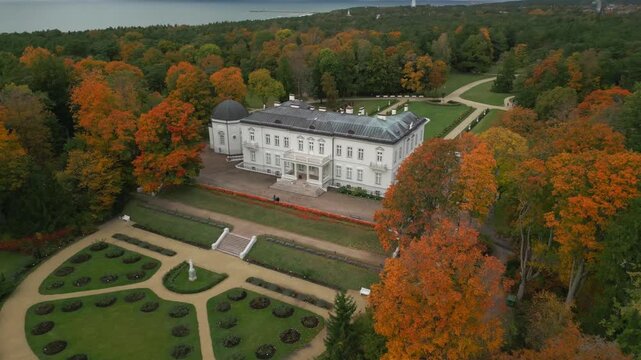 Autumn foliage surrounds a grand white manor and manicured gardens, overlooking a tranquil bay.