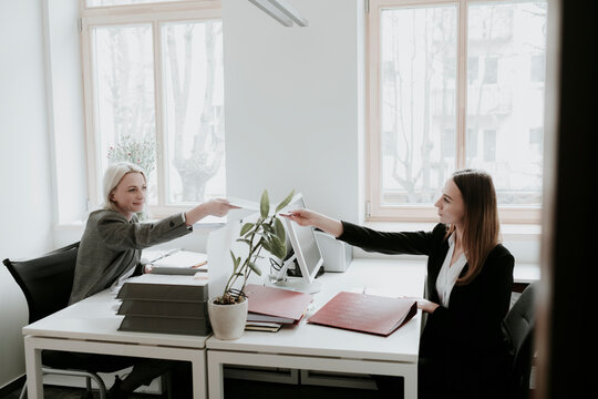 Two young women working at desk in office handing over paper