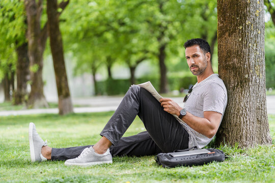 Man leaning against a tree in park reading newspaper