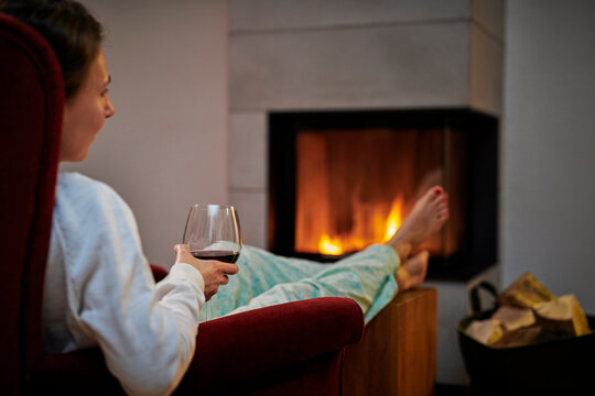 Woman sitting on armchair with glass of red wine relaxing in front of fireplace