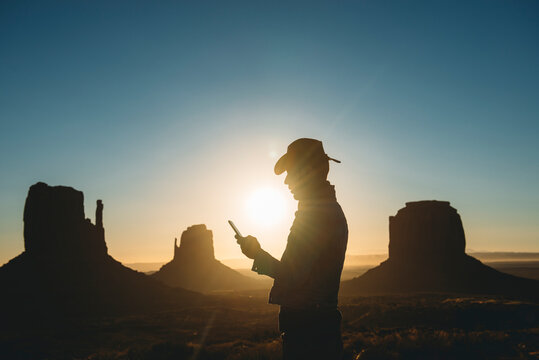 USA, Utah, Monument Valley, silhouette of man with cowboy hat looking at mobile phone at sunrise