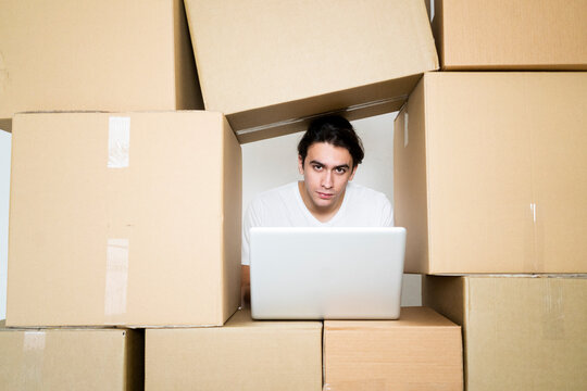 Young man with laptop while trapped between cardboard boxes at new apartment