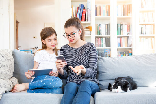 Two sisters sitting on the couch at home using electronic devices