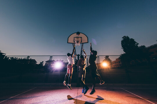 Male African friends playing basketball in court against clear sky at night