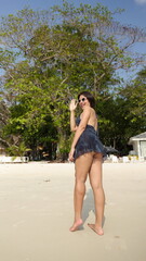 Fototapeta premium A smiling asian woman in a backless blue beach dress and sunglasses waves while walking on a sunny white sand beach
