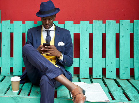 Young businessman wearing old-fashioned suit and hat sitting on a green bench checking his phone