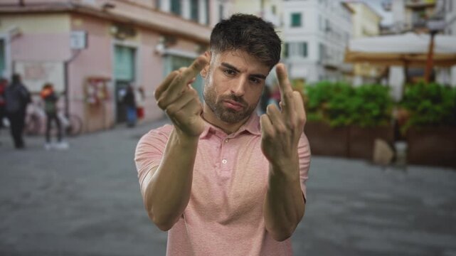 Young hispanic man in pink polo shirt raises middle fingers while walking on urban street; defiance.