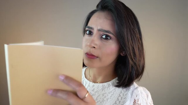 Close up portrait of annoyed Asian student girl young woman holding book looking staring at camera with angry face expression, isolated over grey background.
