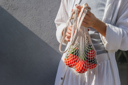 Woman adjusting mesh bag of oranges near textured gray wall in sunlight