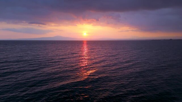 Aerial drone footage of sunset over the Mediterranean Sea in Greece, featuring calm water, bright sun reflection, distant mountains on the horizon, and dramatic evening clouds above the coast.