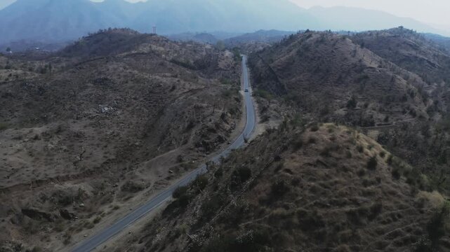 Aerial view of a lone vehicle traveling along a paved road winding through sparse, rocky hills in rural Rajasthan, surrounded by arid scrubland and distant mountain ranges under hazy skies.