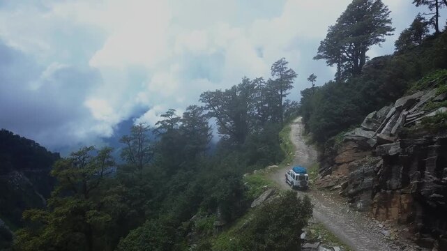 Aerial drone advances from rugged Himalayan mountain road with parked SUV toward dramatic cloud-filled valley in Chamba District, Himachal Pradesh, revealing steep terrain and mist-covered peaks.