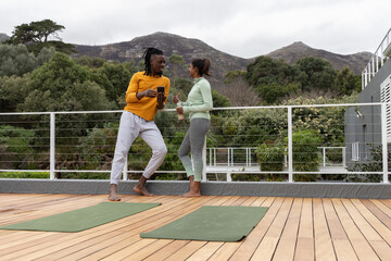 Diverse couple on wood deck with green yoga mats, wearing sportswear holding phone and water bottle © wavebreak3