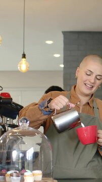 Vertical video: Pouring green-apron barista guiding milk into red cup as customer asking at cafe