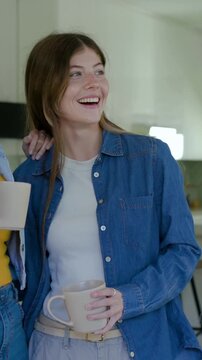 Vertical video: Smiling woman in denim top holding mug after friend placing arm on shoulder at door