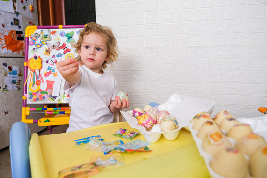Moscow, Russia, 12.04.2025 Blonde child decorating egg for Easter holiday. Little kid holding festive decal near table with prepared ornaments. Creative home activity for childhood development and spr