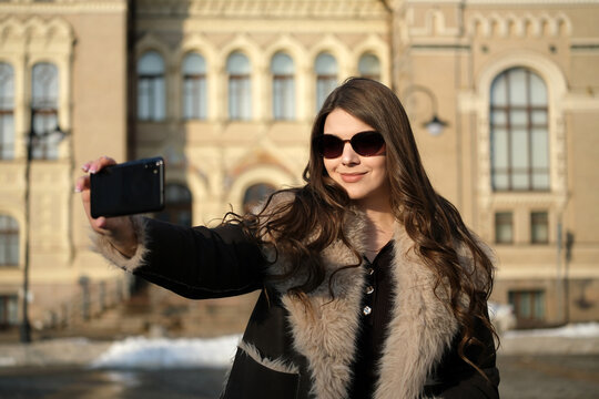 Young woman filming selfie video on smartphone outdoors in early spring sunlight stylish female creating online content social media lifestyle