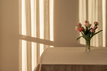 Table with neutral beige linen tablecloth, vase with tulip flowers bouquet, empty wall background with natural warm evening sunlight and shadows, aesthetic minimal floral home interior decor