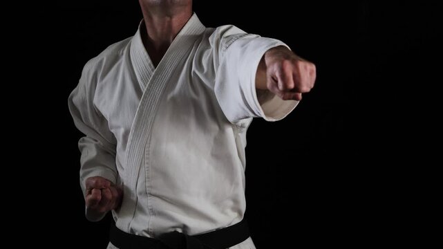 A male athlete with a black belt in karategi practices punches and blocks with his hands on a black background