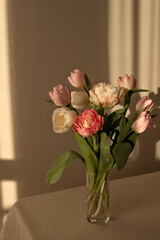 Tulip flowers bouquet in vase on neutral beige linen table cloth, empty wall with aesthetic evening sunlight shadow background, spring or summer home interior decor