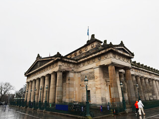 Naklejka premium Neoclassical National Gallery facade with stone columns and flags in Edinburgh, Scotland, UK on a rainy overcast day