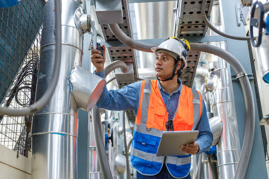 Professional HVAC engineer inspecting industrial ventilation, pipe system in mechanical room of office building. Technician wearing safety helmet using tablet for maintenance, engineering inspection.