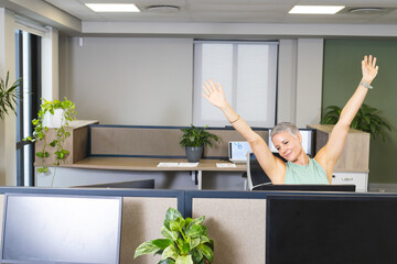 Mature woman in green top stretching arms up in cubicle with plants, copy space © wavebreak3