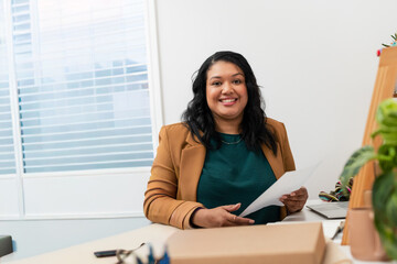 Mid-adult woman sitting at desk in bright workspace, wearing tan blazer, holding document by box © wavebreak3