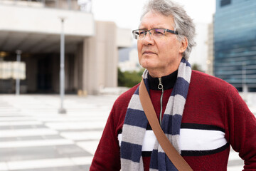 Senior man standing on right in city plaza gazing in red sweater glasses shoulder-strap, copy space © wavebreak3