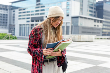Female student standing on rooftop plaza reading green folder and blue notebook, wearing beanie © wavebreak3
