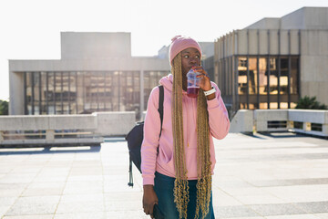 African American woman drinking from bottle in sunlit plaza wearing hoodie, backpack, holding phone © wavebreak3