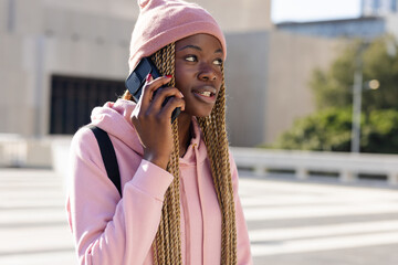 African American woman standing near crosswalk holding phone to ear, wearing pink beanie, hoodie © wavebreak3