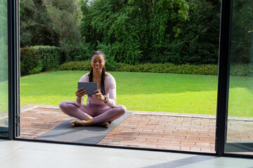 African American woman sitting on patio on yoga mat wearing pink activewear holding tablet © wavebreak3
