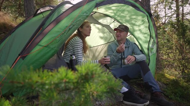 Adult couple talking and drinking tea inside a tent with a scenic coastal view. Authentic camping lifestyle, connection, and slow travel adventure during summer vacation.