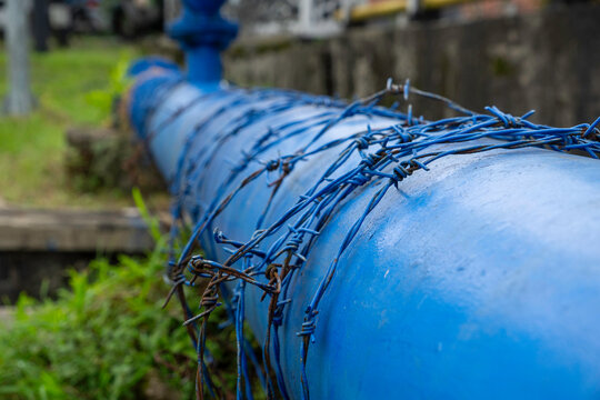 Close-up of a large blue industrial pipe wrapped in blue barbed wire for security. Infrastructure protection in a rural area.