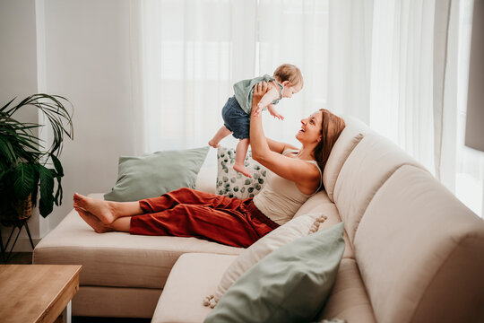 Mother playing and kissing baby on sofa at home in bright living room