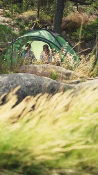 Couple in a green tent among rocks and pine trees in Finland, enjoying peaceful camping in the forest with tall grass in the foreground.