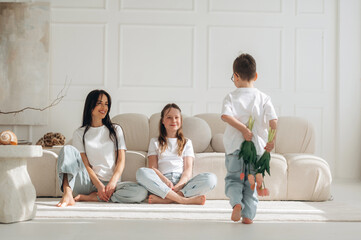 Fototapeta premium Woman and two children sitting on a light-colored rug in a modern living room, child playfully holding green leaves behind back, soft natural light illuminating the scene